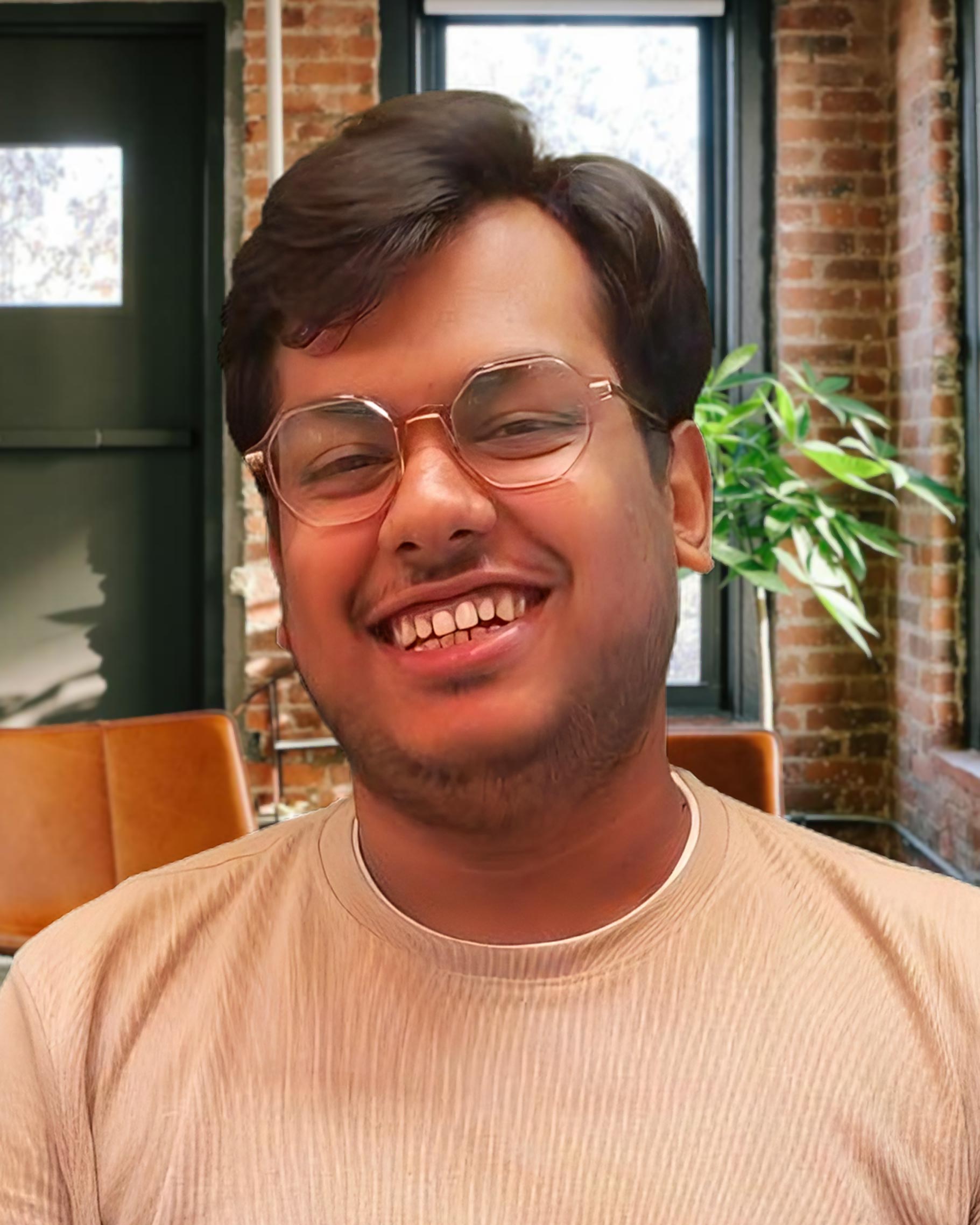 A picture of my where I am glasses and a sporting a subtle beard, and smiling (somewhat, awkwardly though) in a well-lit room with exposed brick walls and large windows. There are green plants and wooden chairs in the background. I am also wearing a beige shirt.